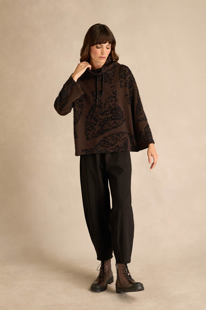 A woman stands against a beige background wearing the Cocoa Speckled Jersey Boxy Top, black loose-fitting pants, and black lace-up boots.