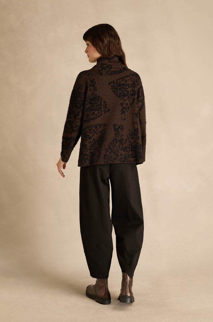 A person stands facing away, wearing the Cocoa Speckled Jersey Boxy Top with a brown abstract pattern and boxy fit, paired with black loose pants and dark ankle boots, against a neutral beige background.