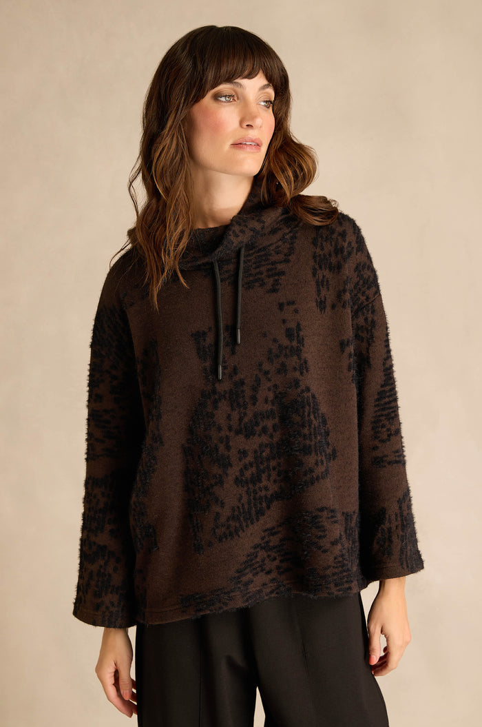A woman with wavy brown hair wears the Cocoa Speckled Jersey Boxy Top—a loose, boxy fit top with a dark brown and black abstract pattern—paired with black pants, standing against a neutral background.