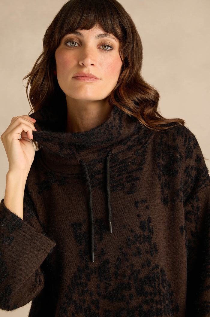 Woman with wavy brown hair wearing the Cocoa Speckled Jersey Boxy Top, a boxy fit featuring a brown and black abstract pattern and cowl-neck with black drawstrings, standing against a neutral background.