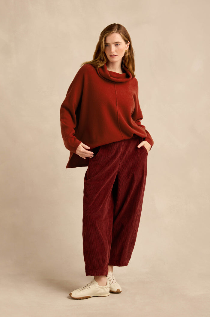 A woman stands against a beige background with her hands in her pockets, wearing a loose red sweater, the Conker Jumbo Cord Bubble Trouser in a wide red silhouette with a comfortable pull-on style, and white sneakers.