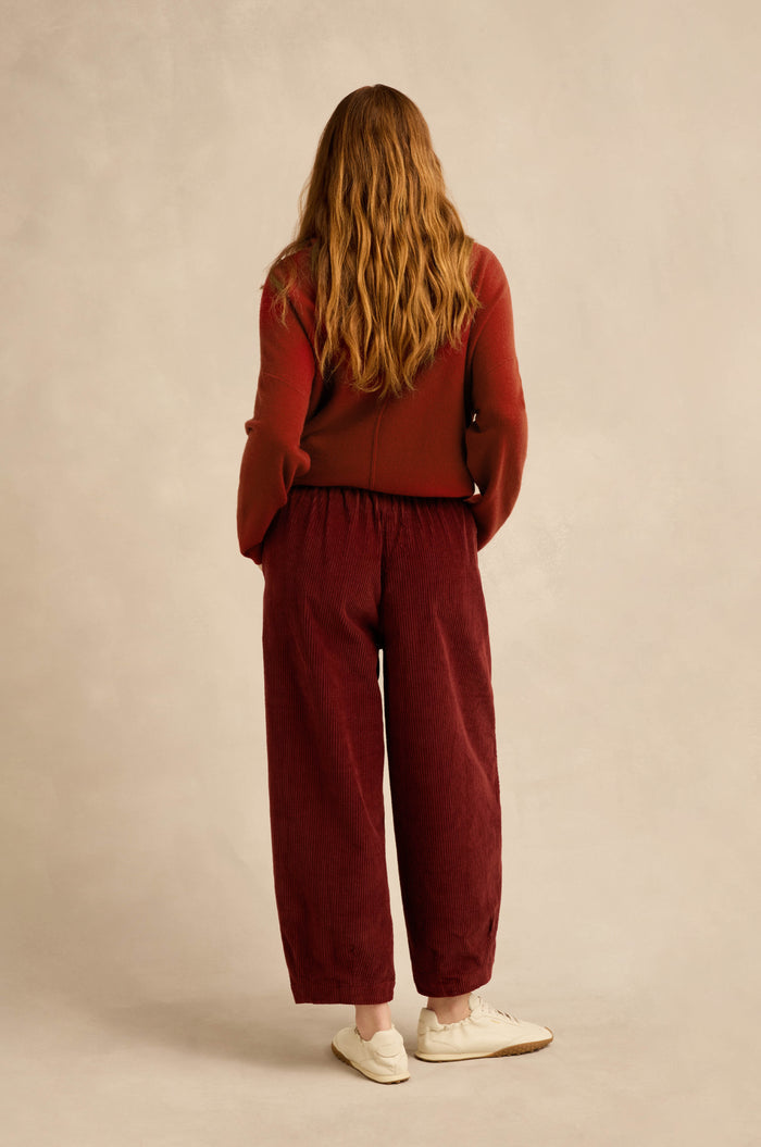 A person with long wavy hair stands facing away, wearing a rust-colored sweater, white sneakers, and Conker Jumbo Cord Bubble Trousers in maroon—a loose pull-on style with a bubble silhouette—against a neutral background.