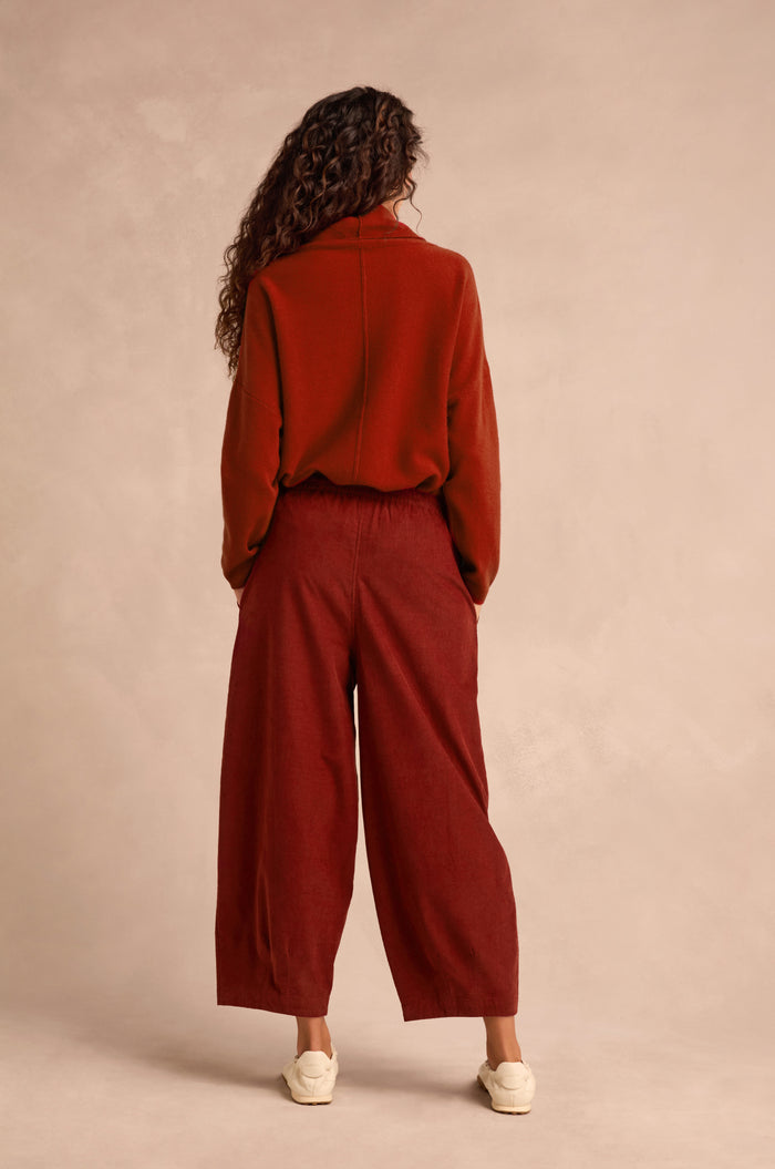 A person with long curly hair stands facing away, wearing relaxed-fit Conker Pinwale Cord Bubble Trousers and a rust-colored fine pincord sweater, paired with white shoes against a neutral background.