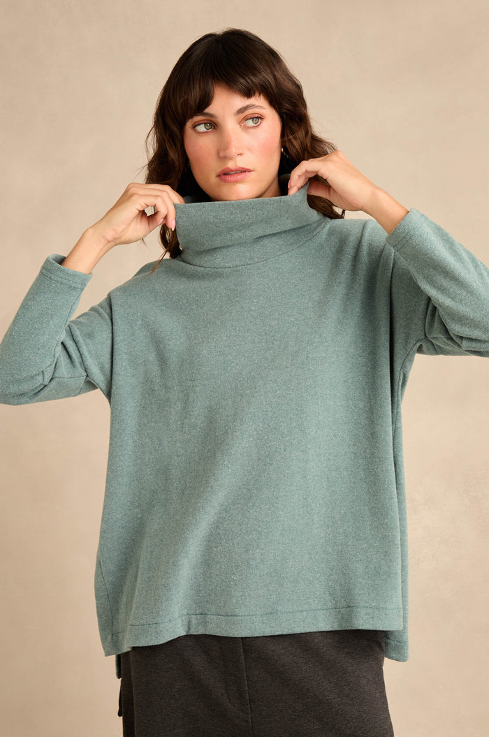 Wearing the Cowl Neck Soft Jersey Top in light teal, this person adjusts the collar of the loose-fitting, cotton-blend top while standing against a neutral background and pairing it with dark pants.