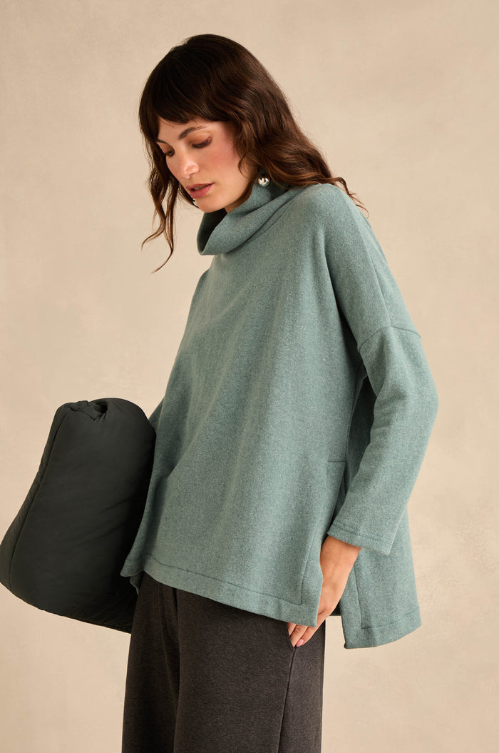A woman wearing the Cowl Neck Soft Jersey Top in light teal paired with dark pants, holding a dark padded object against a beige background.