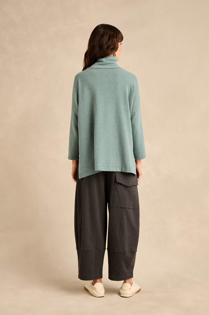 A person stands facing away, wearing the Cowl Neck Soft Jersey Top in a relaxed fit light blue cotton-blend fabric, paired with wide dark gray pants featuring pockets and beige slip-on shoes against a neutral background.
