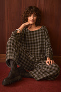 A person with curly hair sits on a red carpet, styled in the Crinkle Check Boxy Top and black boots, posed against a wooden wall.