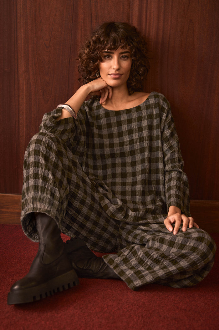 A person with curly hair sits on a red carpet, styled in the Crinkle Check Boxy Top and black boots, posed against a wooden wall.