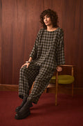 A person with curly hair sits on a wooden chair in a room with wood-paneled walls and red carpet, wearing Crinkle Check Bubble Trousers with a cropped leg and black platform boots.