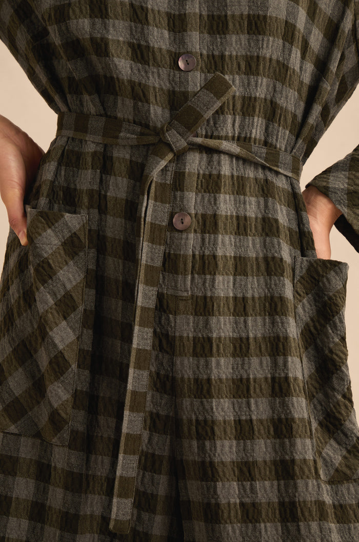 Close-up of a person wearing the Crinkle Check Flared Jumpsuit, which features a textured plaid button-down design, matching belt tied at the waist, and hands in front patch pockets.
