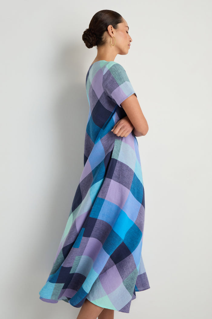 Woman in profile wears the Cross Dye Check Linen Maxi Dress, a flowing short-sleeve v-neck style with large blue, purple, and teal checkered pattern, standing against a plain white background.