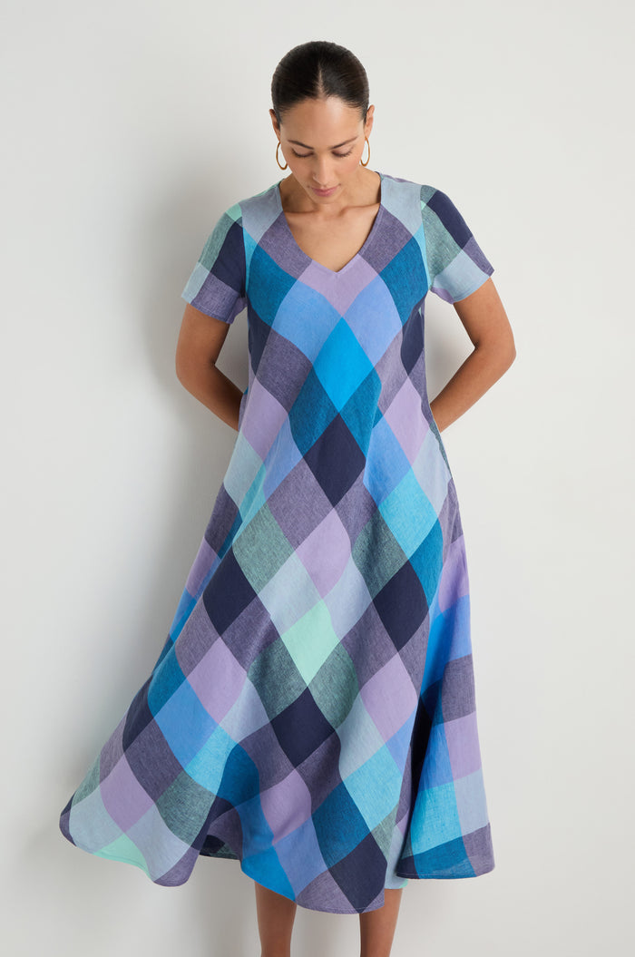 A person wears the Cross Dye Check Linen Maxi Dress, a short-sleeve V-neck maxi in blue, purple, and teal checkered linen, standing against a plain white background.
