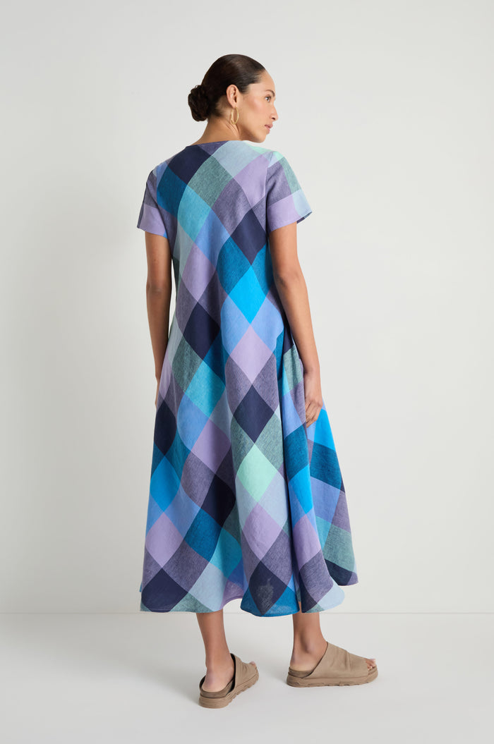 A woman faces away, wearing the Cross Dye Check Linen Maxi Dress in blue and purple tones with beige sandals, set against a plain white background.