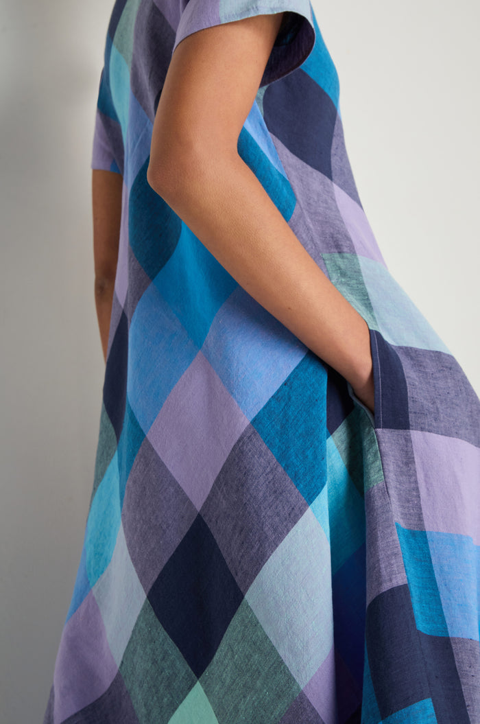 A person stands against a plain background with one hand in their pocket, wearing the Cross Dye Check Linen Maxi Dress—featuring short sleeves and large blue, purple, and teal check patterns.