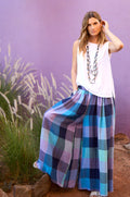 A woman stands outdoors against a purple wall, wearing a white sleeveless top, Cross Dye Check Linen Wide Trousers in blue and purple tones, and layered necklaces.