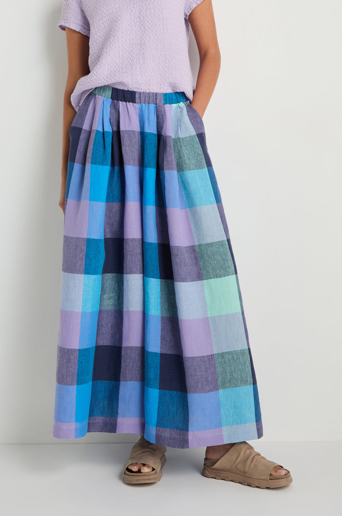 A person wearing a light purple short-sleeve top, Cross Dye Check Linen Wide Trousers with an ultra wide leg in blue and purple checks, and tan sandals stands against a plain light-colored wall.