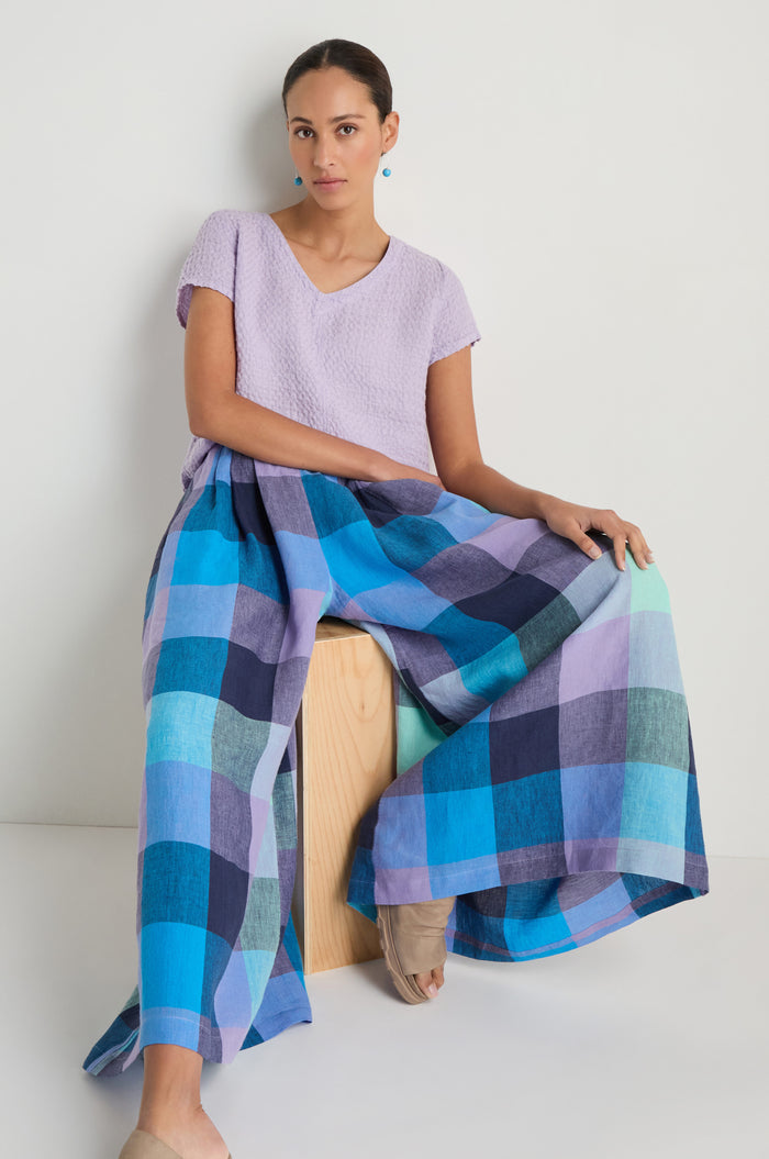 A woman sits on a wooden block, wearing a light purple textured top and blue and purple Cross Dye Check Linen Wide Trousers, against a plain white background.
