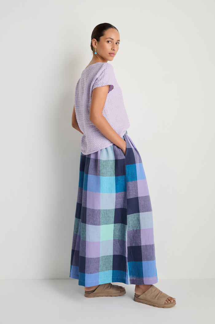 A woman stands against a white wall wearing a short-sleeve textured lavender top, Cross Dye Check Linen Wide Trousers in blue and purple plaid, tan sandals, and green earrings.