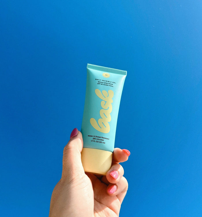 Hand holding a blue bask sunscreen tube with yellow label against a solid blue background.