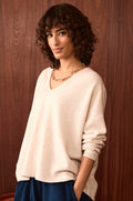 A woman with curly hair wears the Ecru Merino Wool V Neck Jumper, blue pants, and a gold chain necklace as she stands in front of a wooden wall, looking at the camera with a slight smile.