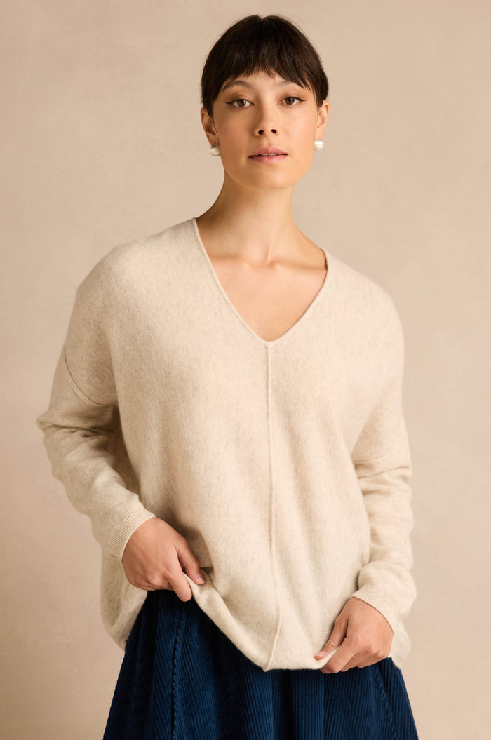 A woman wearing an Ecru Merino Wool V Neck Jumper and a navy pleated skirt stands against a beige background, looking at the camera.