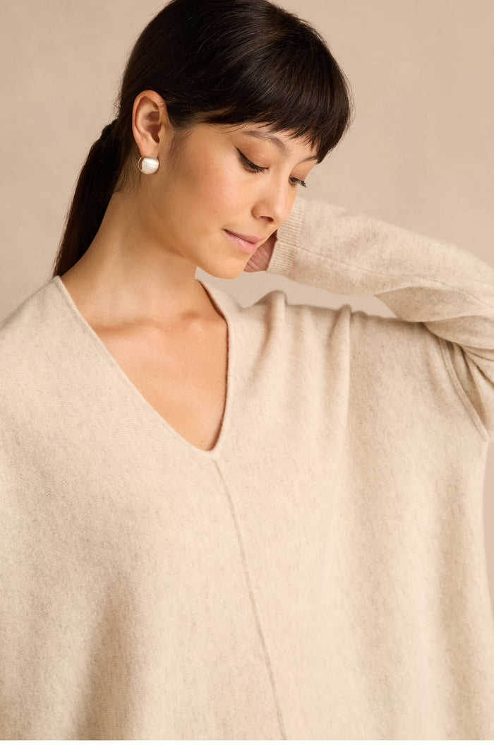 A woman with dark hair and pearl earrings wears an Ecru Merino Wool V Neck Jumper, looking down with one hand gently touching the back of her neck.