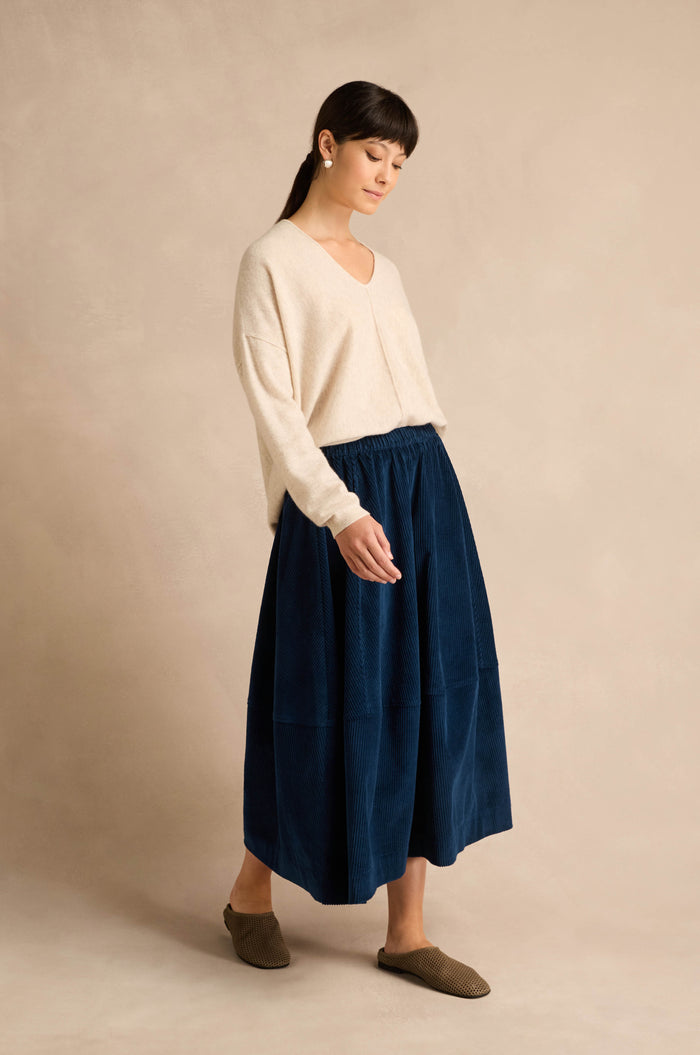 A woman wearing an ecru Merino Wool V Neck Jumper, a dark blue midi skirt, and brown slip-on shoes stands against a plain beige background, looking downward.