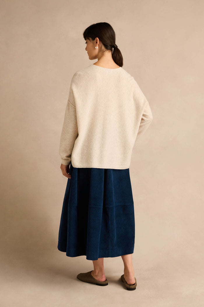A woman stands facing away, wearing the Ecru Merino Wool V Neck Jumper, a blue midi skirt, and brown slip-on shoes, against a neutral beige background.