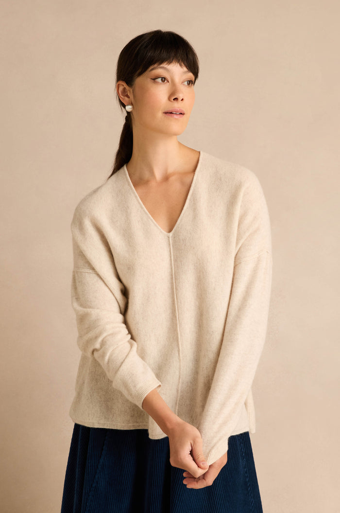 A woman wearing the Ecru Merino Wool V Neck Jumper and a blue skirt stands against a beige background, looking to the side.