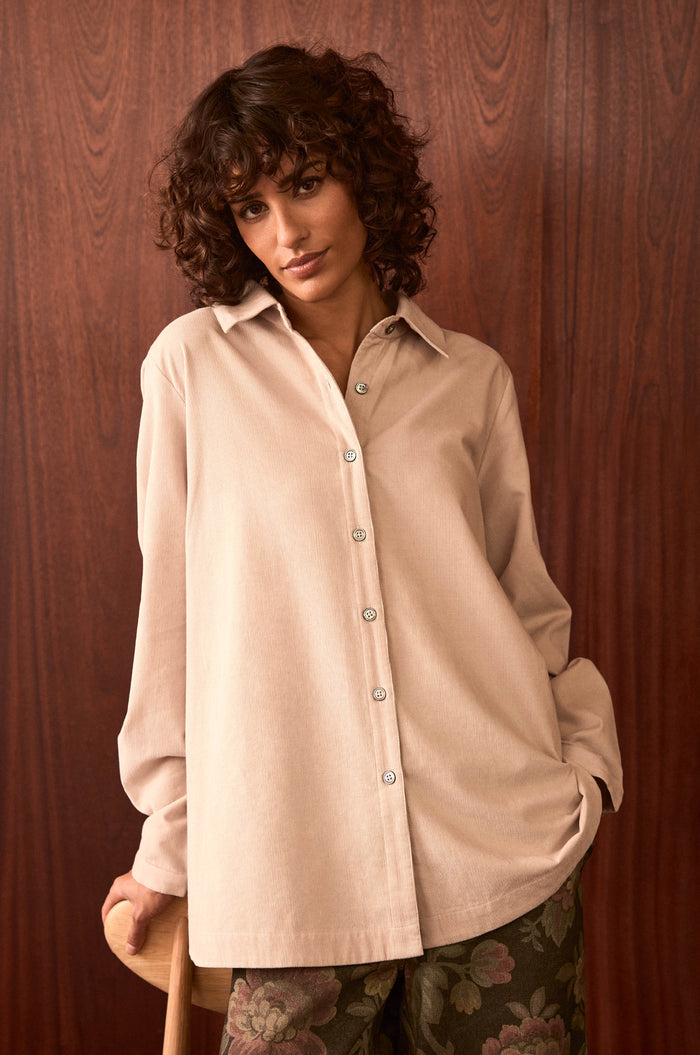 Woman with short curly hair wearing an Ecru Pinwale Cord Shirt and patterned pants stands in front of a wooden wall, resting one hand on a chair.