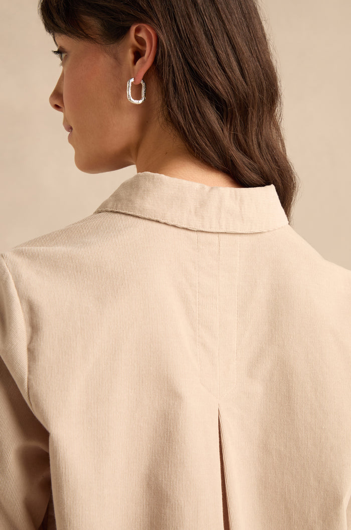 A woman with long brown hair wears the Pinwale Cord Shirt in beige, featuring a pure cotton fabric and a collared neckline, paired with a silver hoop earring; she is shown from behind and in profile against a neutral background.