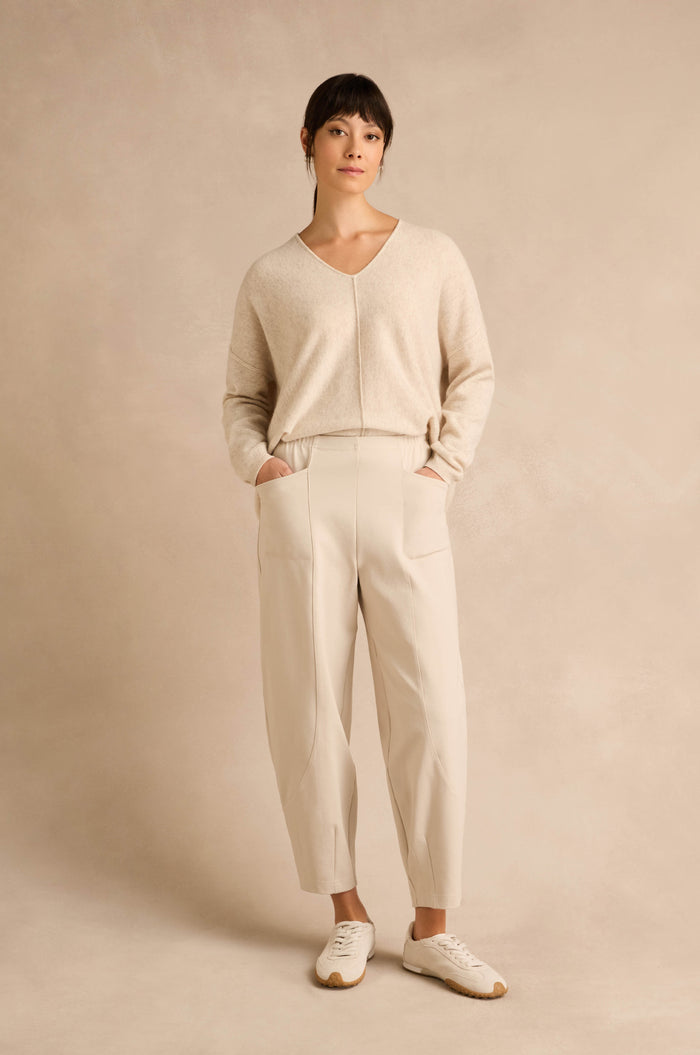 A woman stands against a beige background, wearing a light beige sweater, Ecru Ponte Bubble Trousers in a cream-colored hue that create a subtle bubble silhouette, and white sneakers, with her hands in her pockets.