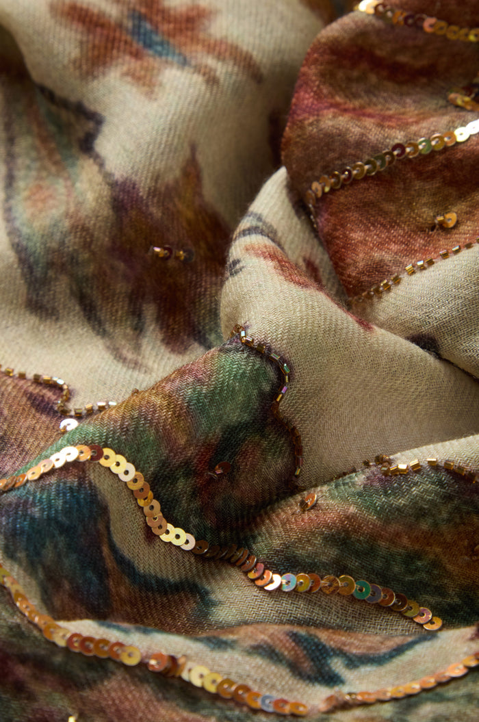 Close-up of the Embroidered Tapestry Wool Scarf, showcasing beige wool fabric adorned with a multicolored pattern and gold sequin embroidery in wavy lines, creating a richly textured and shimmering surface.