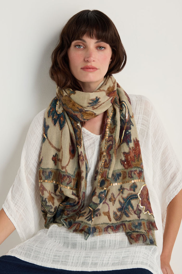 A woman with brown hair wears a white textured top and the Embroidered Tapestry Wool Scarf, featuring a beige base with a delicate floral pattern, sitting against a plain light background.