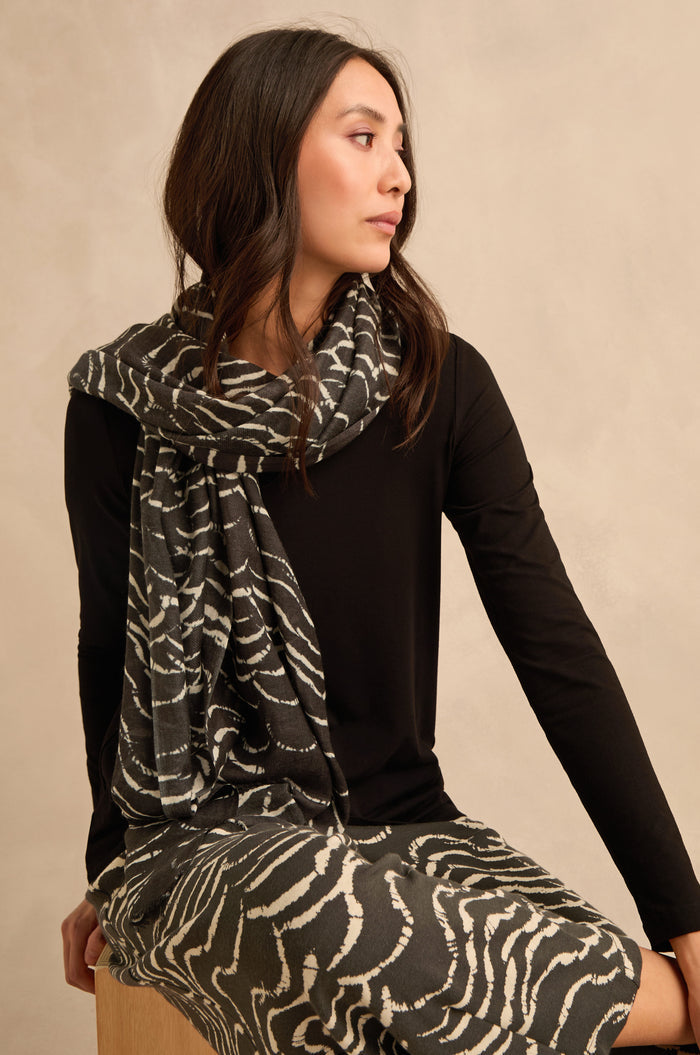 A woman sits on a wooden stool, wearing a black long-sleeve shirt and the Dancing Waves Scarf—a lightweight winter accessory featuring a striking wave pattern—paired with a matching skirt, looking to the side against a beige background.