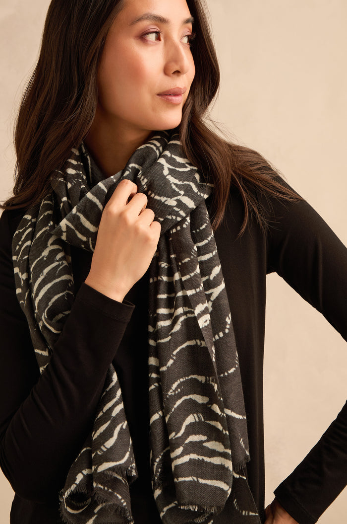 A woman wearing a black long-sleeve top drapes the Dancing Waves Scarf, featuring a striking black-and-white wave pattern, and gazes to the side while holding the scarf.