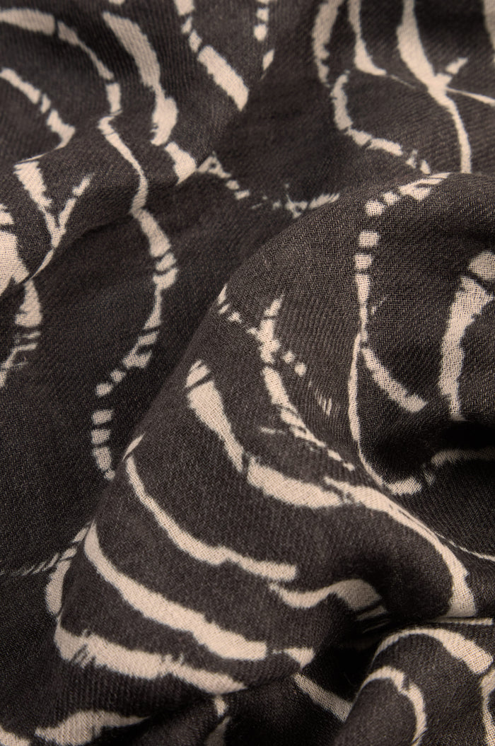 Close-up of the Dancing Waves Scarf, crafted from a wool-silk blend in black and adorned with a white, abstract wavy line pattern. This lightweight winter accessory showcases visible folds and a rich texture.
