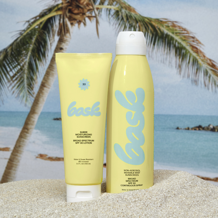 Two yellow bask sunscreen containers (tube and spray) on sand with palm fronds and ocean in the background