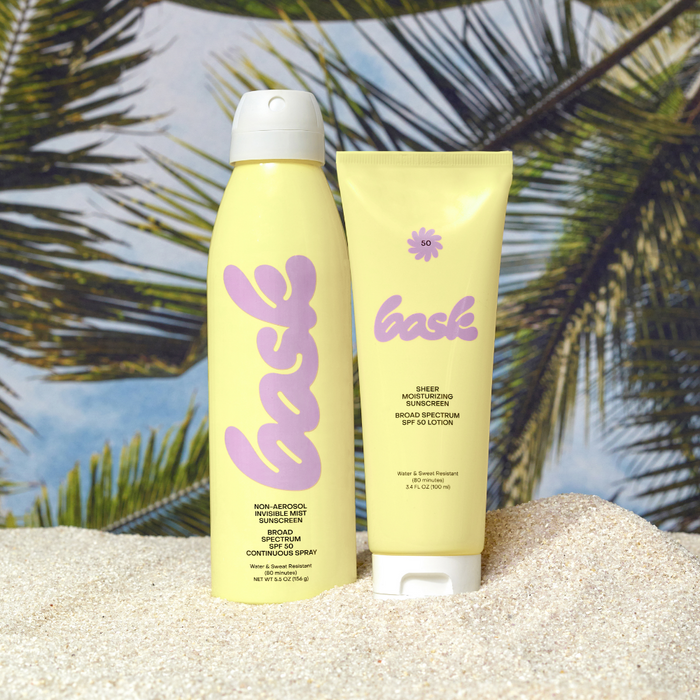 Two yellow sunscreen tubes with pink script bask logo on a sandy beach and palm trees in background.
