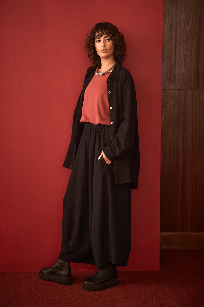 A woman with curly hair stands against a red wall, wearing a red top, black Fine Crinkle Barrel Trousers in a cropped length, a black jacket, and black platform shoes on a red carpet.