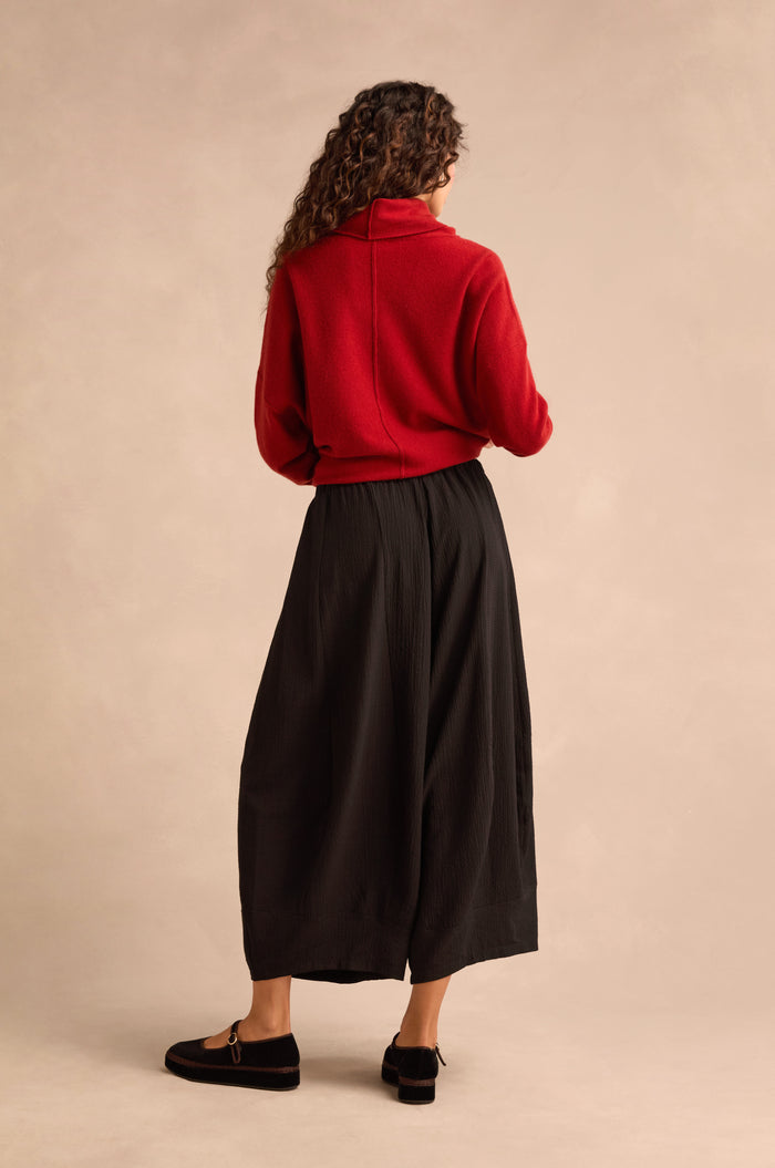 A person is standing with their back turned, wearing a red turtleneck sweater, Fine Crinkle Barrel Trousers in black with a cropped length, and black loafers, set against a neutral studio background.
