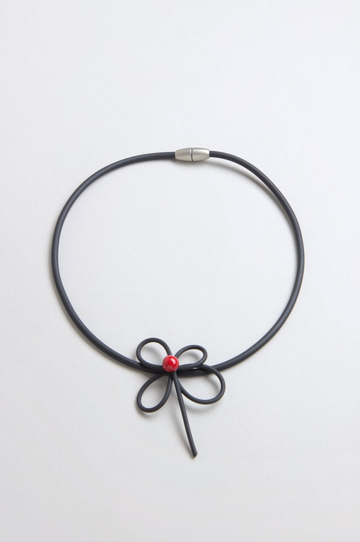 The Flower Necklace features a bow-shaped center with a single red bead, reminiscent of handmade ceramic jewelry, displayed against a plain light background.