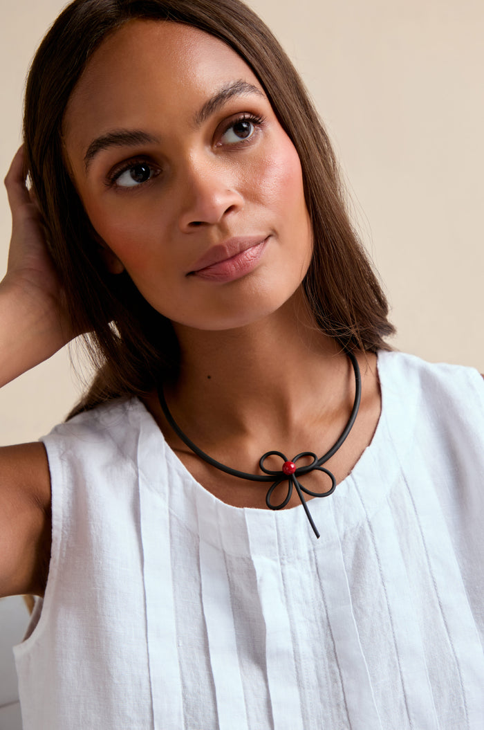 A woman in a white sleeveless top wears the handmade Flower Necklace featuring a bow-shaped pendant and red bead, looking slightly upward with one hand behind her head.