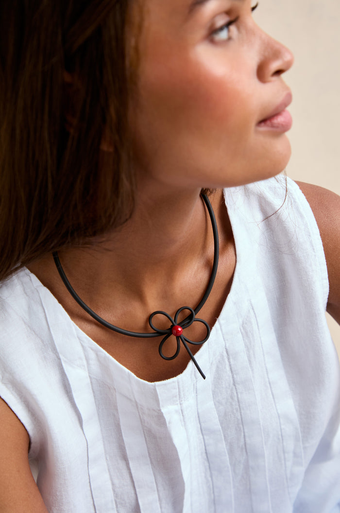 A woman in a white sleeveless top wears the Flower Necklace, a black cord handmade ceramic flower pendant with a red bead, while looking to the side.
