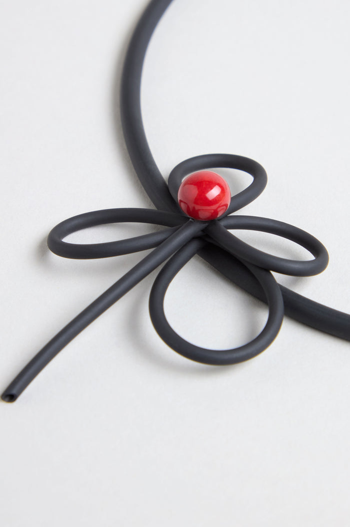 The Flower Necklace features a black flexible cord shaped like a flower with a shiny red bead at the center, displayed on a plain light background.
