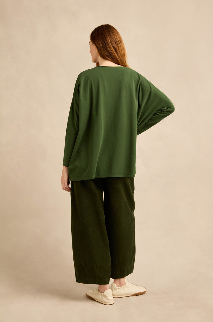 A person is seen from behind, wearing the Forest Cotton Jersey Pocket Top in a loose, boxy silhouette with wide-leg dark green pants and white shoes, set against a neutral beige backdrop.