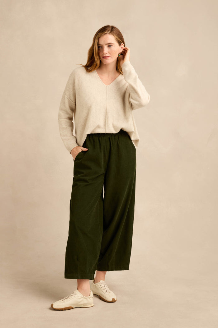 A woman stands against a beige background wearing a beige sweater, the Forest Pinwale Cord Bubble Trousers in a relaxed dark green fit, and white sneakers, with one hand in her pocket and the other touching her hair.