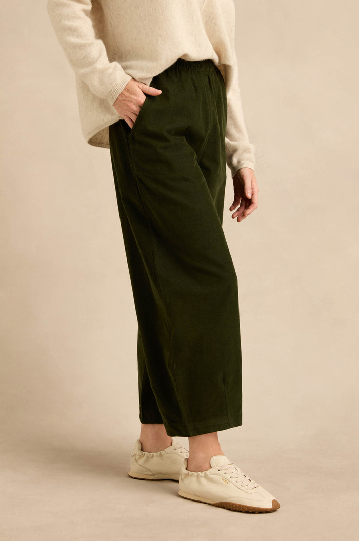 A person wearing a cream sweater, Forest Pinwale Cord Bubble Trousers in dark green, and cream sneakers stands with one hand in their pocket against a beige background.