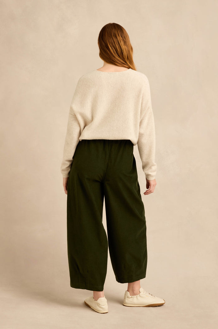 A person stands with their back to the camera, wearing a cream sweater and the Forest Pinwale Cord Bubble Trousers in dark green, featuring a relaxed fit, paired with white sneakers against a plain beige background.