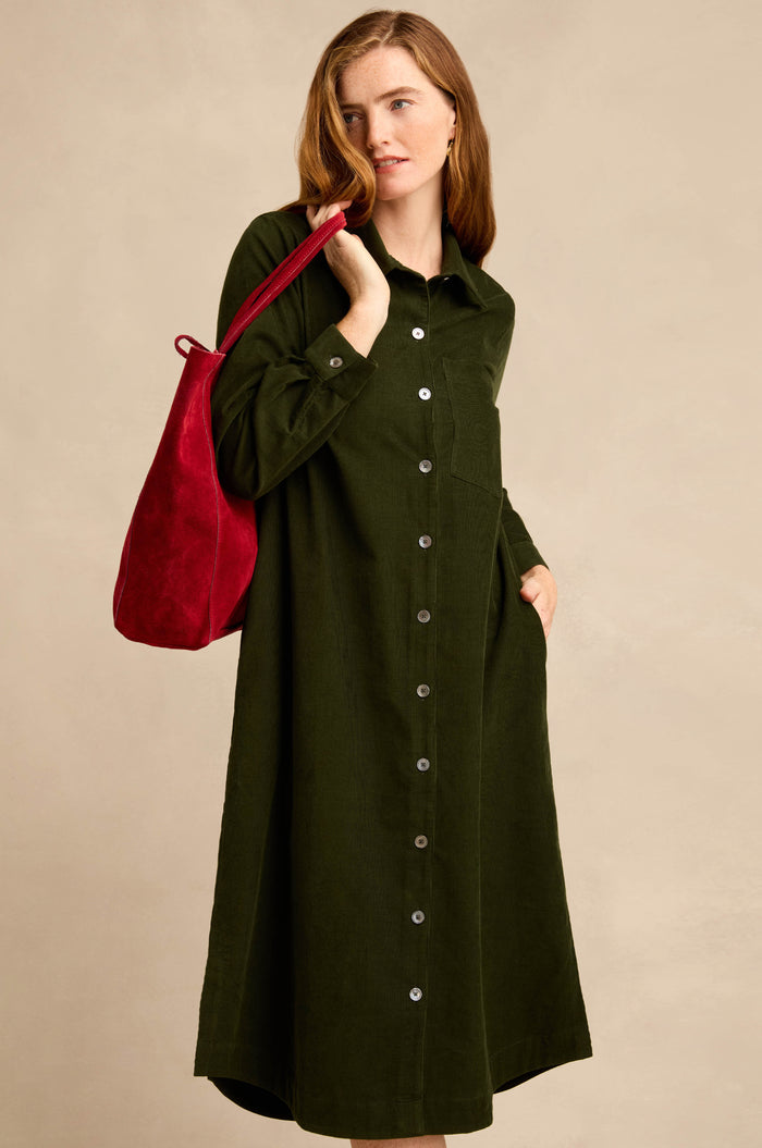 A woman stands against a plain beige background, wearing the Forest Pinwale Cord Shirt Dress—a long-sleeve, button-down, dark green corduroy dress with a breast pocket—while carrying a red shoulder bag.